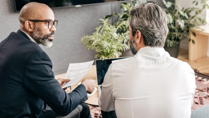 deux hommes discutant l'un à côté de l'autre en tenue professionnelle, de dos, l'un avec une feuille dans la main, l'autre devant un ordinateur