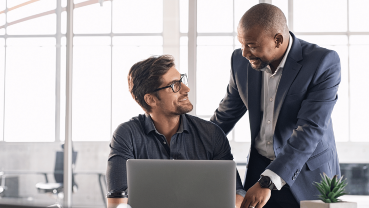 Deux hommes en tenue de travail. L'un est assis devant son ordinateur. L'autre est debout à côté de lui. Ils discutent et se sourient.